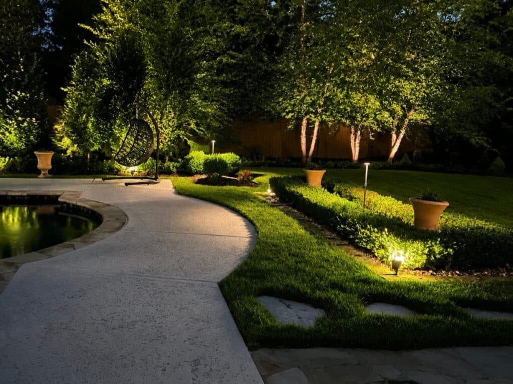 Backyard landscape at night illuminated by low-voltage landscape lighting, highlighting a curved paved path, manicured lawn and hedges, trees, and a pool edge with a hanging egg chair seating area.