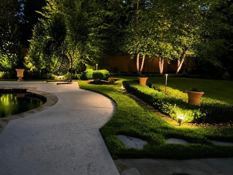 Backyard landscape at night illuminated by low-voltage landscape lighting, highlighting a curved paved path, manicured lawn and hedges, trees, and a pool edge with a hanging egg chair seating area.