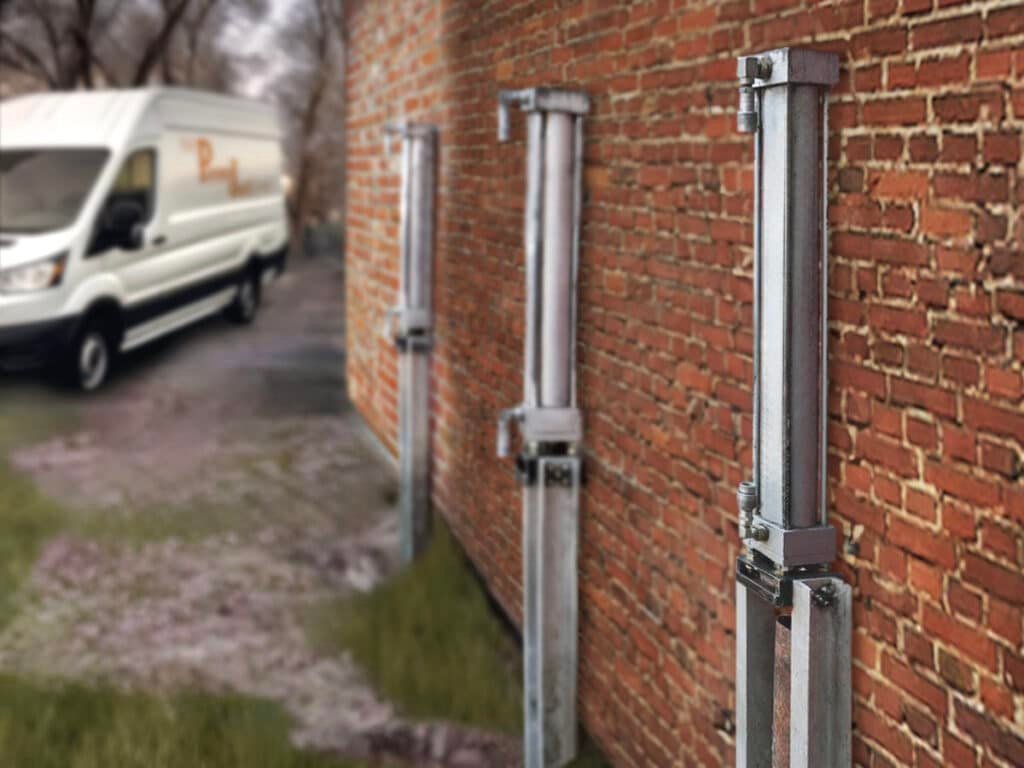 Steel foundation piers installed along an exterior brick wall, supporting structural repair work, with a company service van visible in the background.