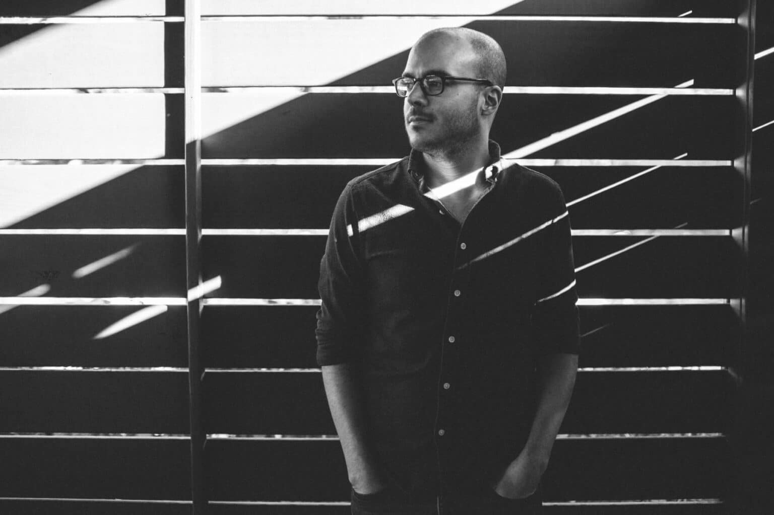 Black-and-white portrait of a man with glasses standing against a striped, light-and-shadow backdrop, looking off to the side.