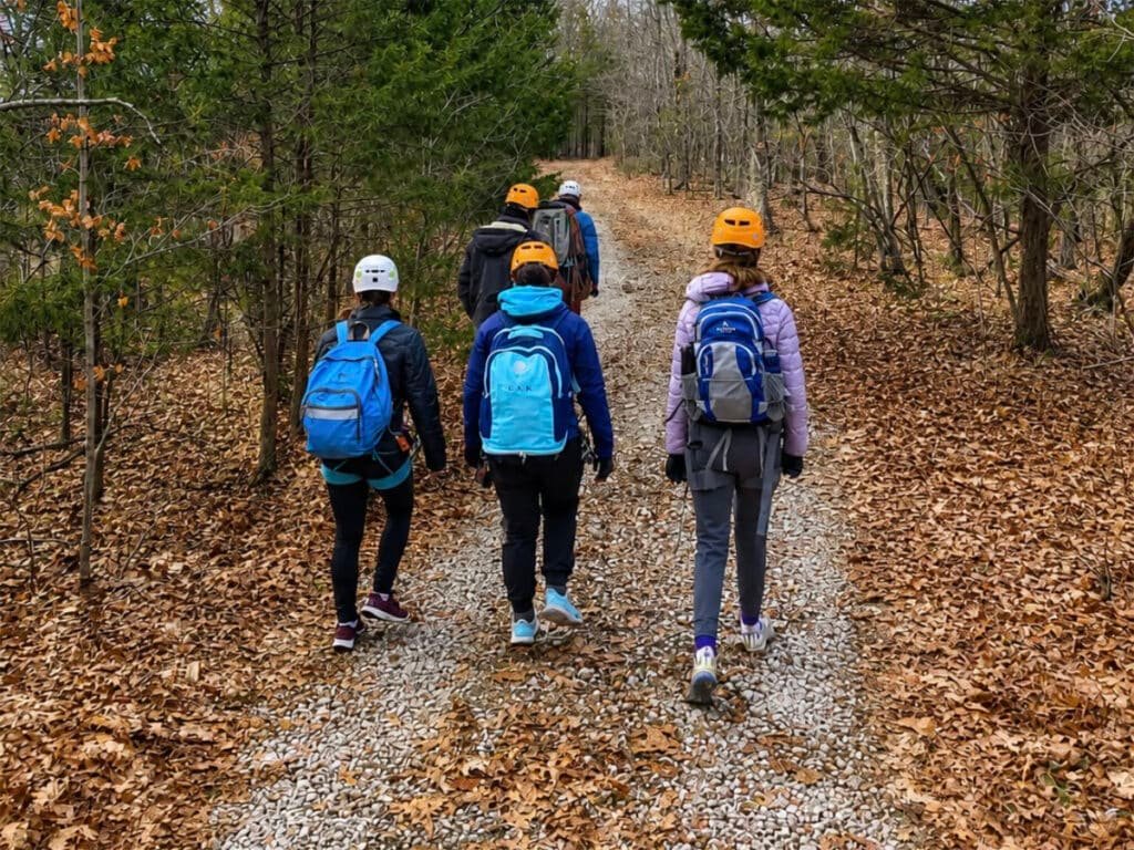 A group of hikers wearing helmets and backpacks walks along a gravel trail through a wooded autumn landscape, viewed from behind.
