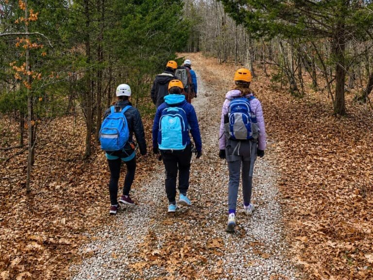 A group of hikers wearing helmets and backpacks walks along a gravel trail through a wooded autumn landscape, viewed from behind.