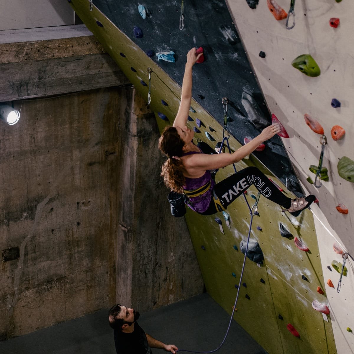 A climber in a purple top and black leggings reading "Take Hold" navigates an overhanging climbing wall, reaching for a red hold while clipped into a rope. A belayer below watches and manages the rope. The gym features green, black, and white climbing surfaces with colorful holds and exposed concrete walls.