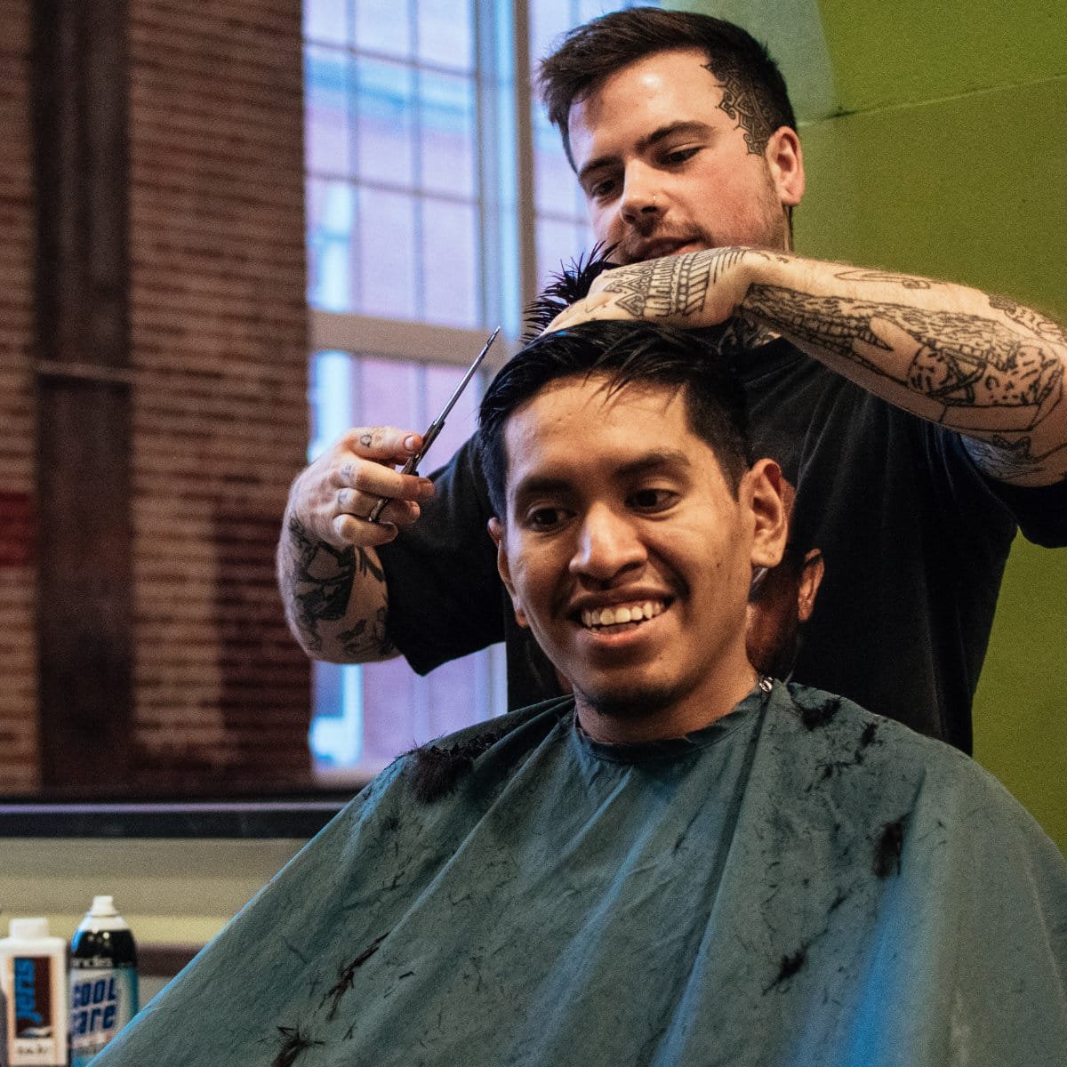 A tattooed barber cuts a smiling client's hair with scissors and a comb in a barbershop. The client wears a gray cutting cape, and hair clippings are scattered across it. Exposed brick, a large window, and green-painted walls are visible in the background, along with barbershop products on the counter.