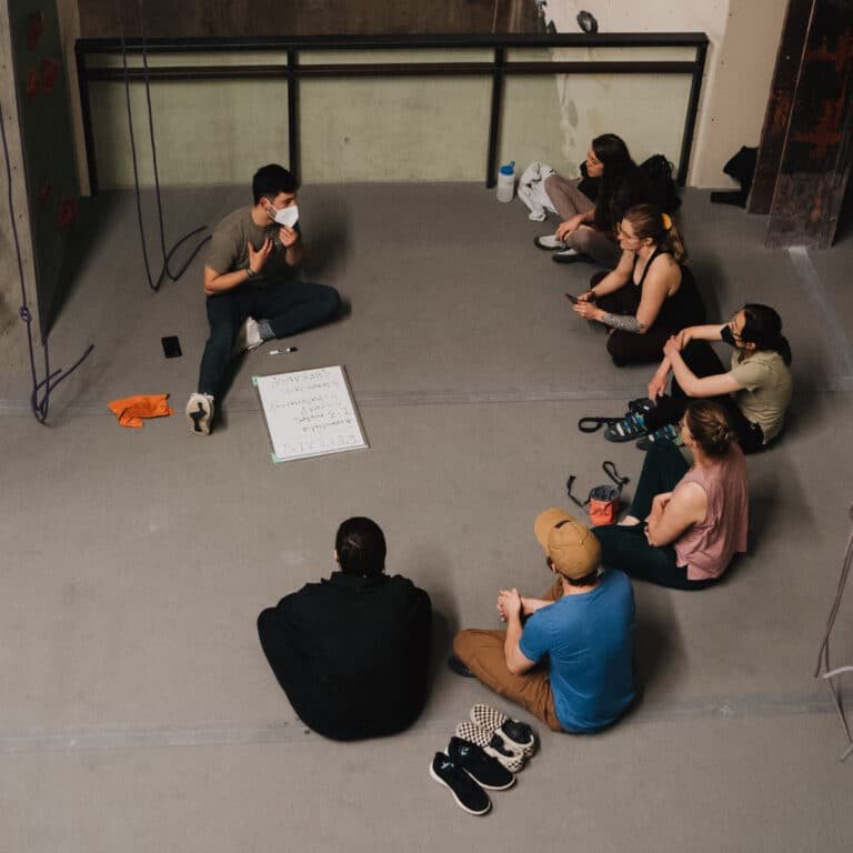 A group sits cross-legged in a circle on the floor of a climbing gym for a discussion, gathered around a small whiteboard with handwritten notes. One person wearing a mask speaks while gesturing, and the others listen. Climbing ropes hang against a wall in the background, and shoes are set aside on the floor.