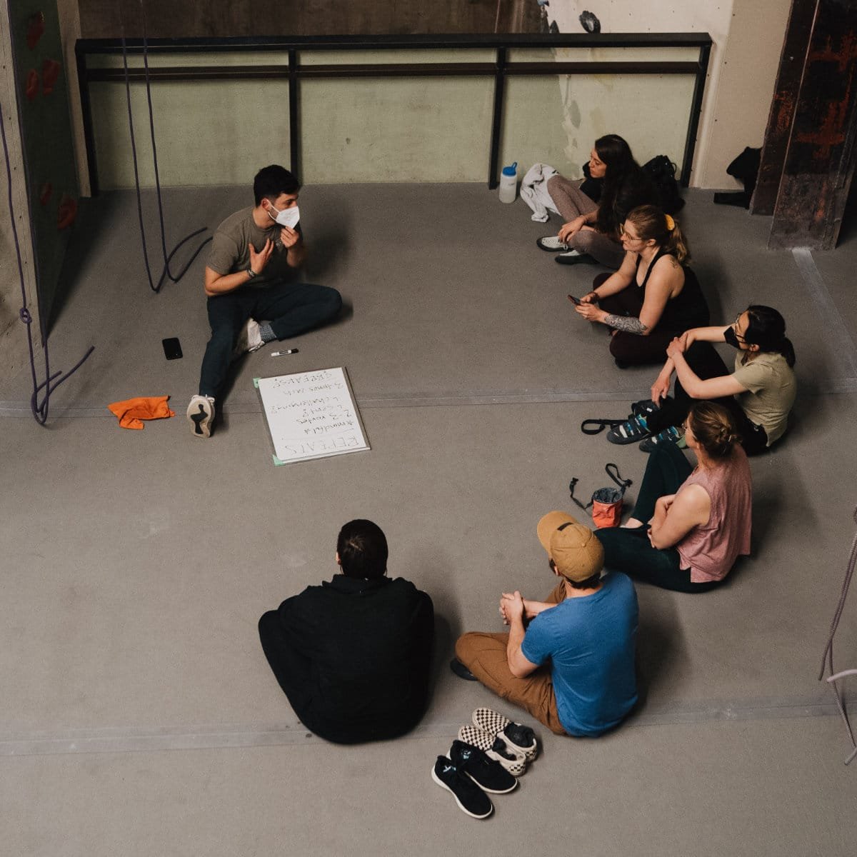 A group sits cross-legged in a circle on the floor of a climbing gym for a discussion, gathered around a small whiteboard with handwritten notes. One person wearing a mask speaks while gesturing, and the others listen. Climbing ropes hang against a wall in the background, and shoes are set aside on the floor.