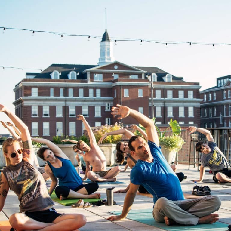 A group practices a seated side-bend yoga stretch on a rooftop, arms raised overhead on colorful mats. A brick building with a white cupola and string lights overhead form the backdrop under a clear sky.