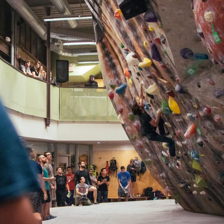 A climber ascends a large overhanging boulder wall covered in colorful holds while a crowd watches from the floor below and a balcony above, in a gym with exposed ductwork and a DJ station off to the side, suggesting a climbing competition or event.