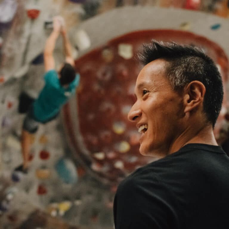 A man with a spiked hairstyle smiles while looking off to the side in a climbing gym, with a climber ascending an overhanging red-toned wall blurred in the background.