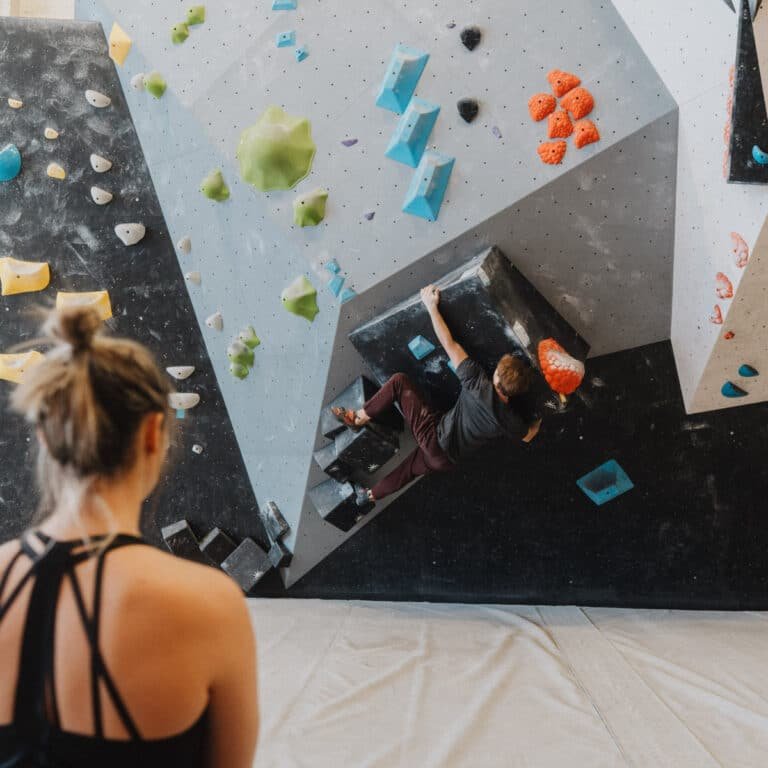 A woman watches a climber navigate an angular bouldering wall with black, gray, and white panels dotted with blue, green, orange, and yellow holds. The climber reaches for a hold on an overhanging volume feature while padding covers the floor below.