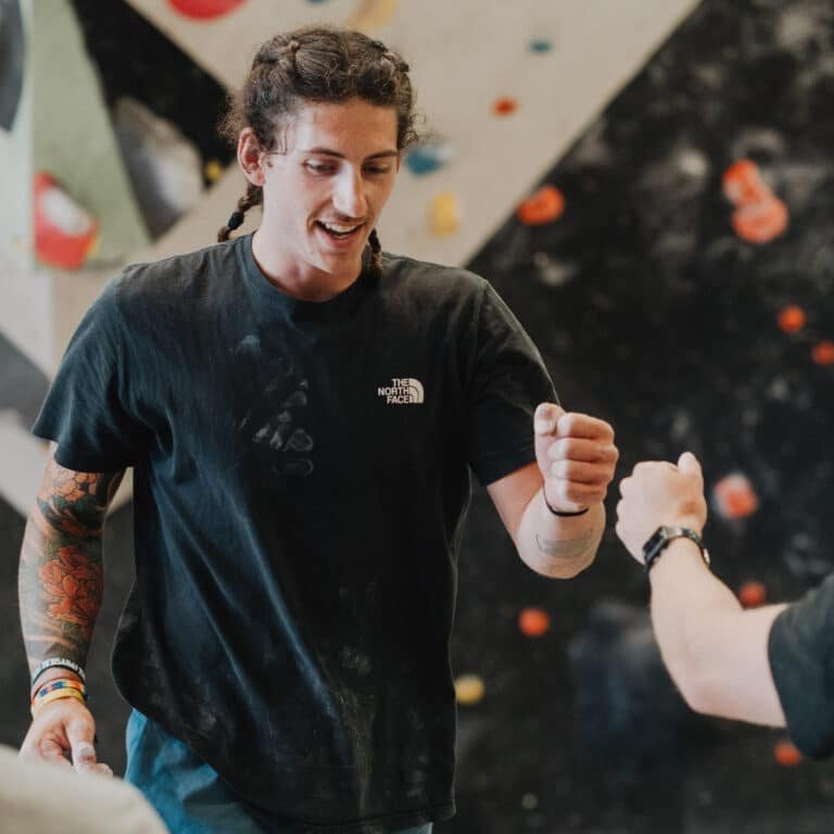 Fist bump between two climbers at Climb So iLL bouldering gym, colorful climbing holds on the wall behind them.