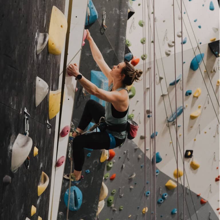 A woman climbs a roped route on a gray wall studded with yellow and white holds, reaching upward with her chalk bag at her hip. Multiple hanging ropes and a colorful wall of holds are visible in the background of the climbing gym.