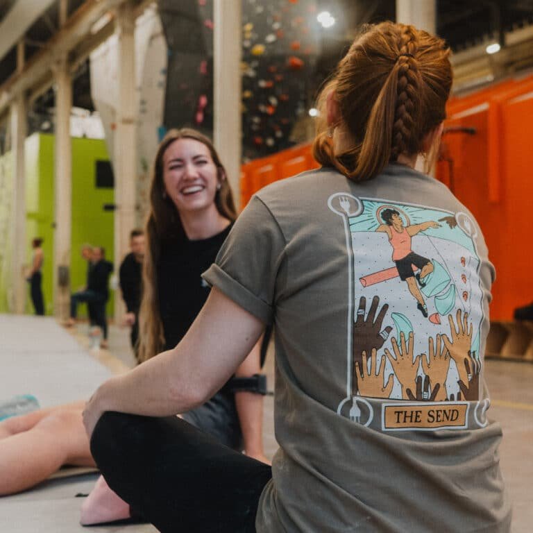 A woman with a braided ponytail sits with her back to the camera, wearing a gray T-shirt with a tarot-card-style graphic and the caption "The Send." Another woman laughs beside her in a climbing gym with an orange bouldering wall visible in the background.