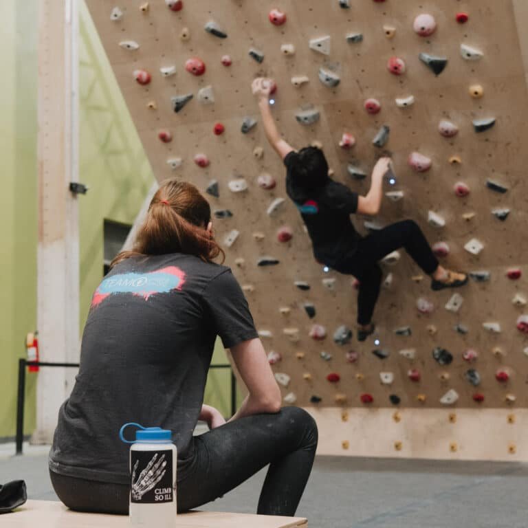 A person wearing a "Team Climb So iLL" T-shirt watches a climber ascend a wood-paneled wall covered in pink, gray, and tan holds. A water bottle with a skeleton hand graphic and "Climb So iLL" branding sits on the bench in the foreground.