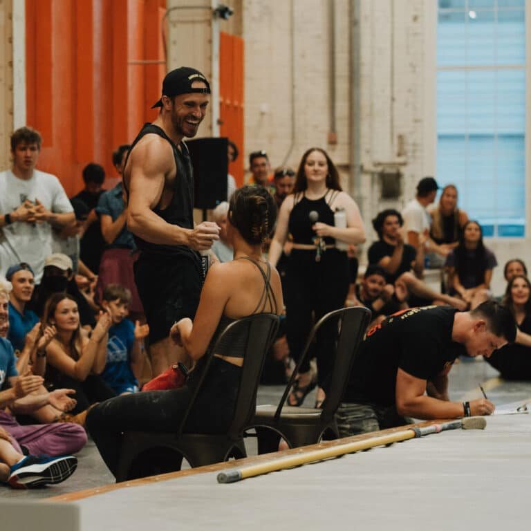 A man in a black tank top smiles and fist-bumps a seated woman during what appears to be an award ceremony or event at a climbing gym, as a crowd sits and applauds on the floor around them. Someone signs paperwork in the foreground while another person holds a microphone in the background.