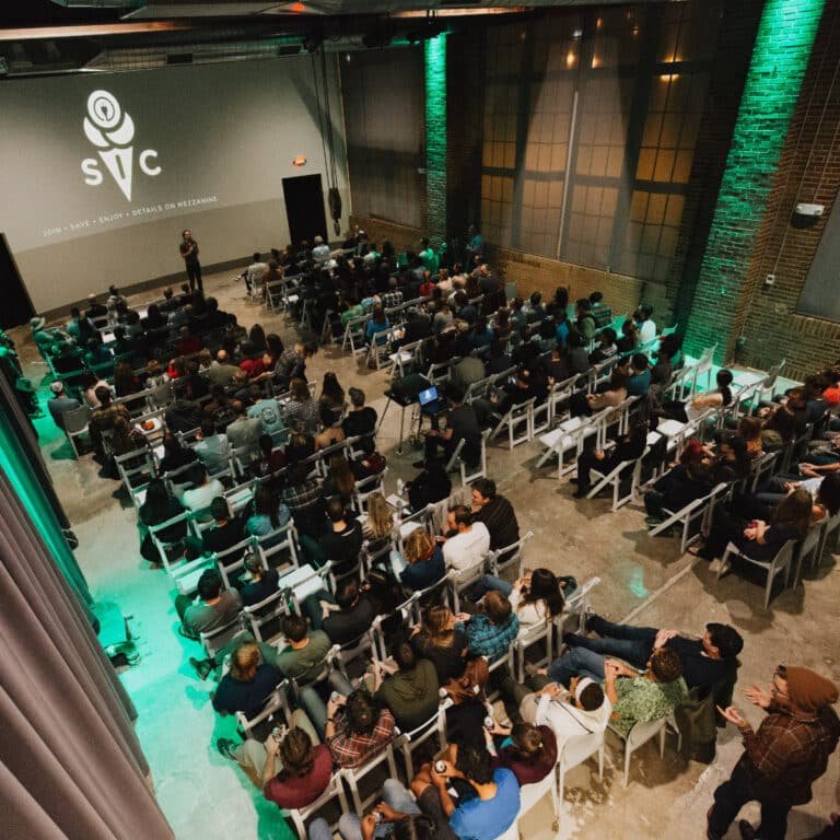 An aerial view of a large seated crowd in a brick-walled venue lit with green ambient lighting, facing a screen displaying an "SIC" logo with membership program text. A speaker stands at the front near the projection screen addressing the audience.