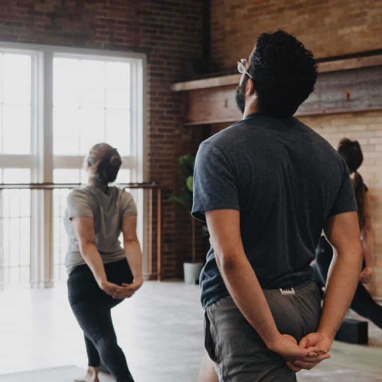 A man clasps his hands behind his back while looking upward during a stretching or yoga session, with others doing similar poses in a bright studio with exposed brick walls and large windows.