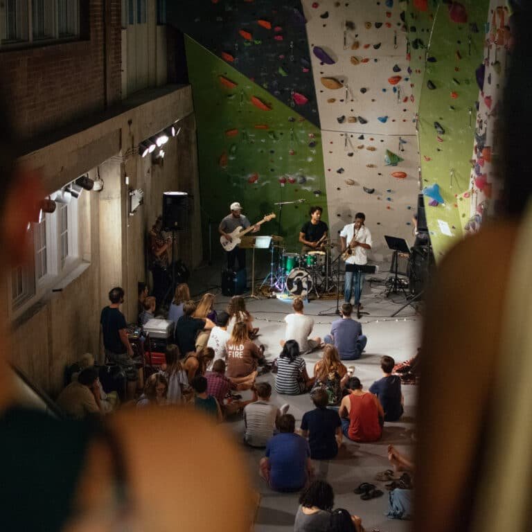 A live band performs with guitar, drums, and saxophone at the base of a green and black bouldering wall, while a seated crowd listens on the padded floor of a climbing gym.