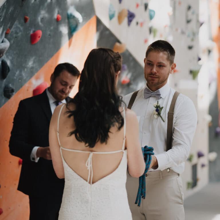 A groom in suspenders and a bow tie holds a coiled climbing rope while facing his bride during a wedding ceremony, with an officiant looking on. The scene takes place in front of a colorful bouldering wall inside a climbing gym.