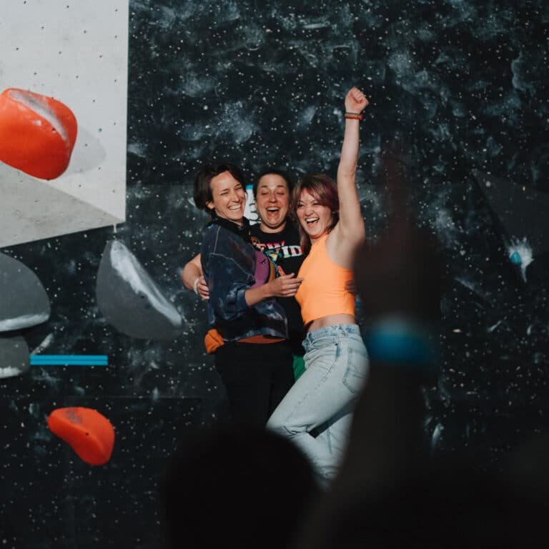 Three women embrace and celebrate joyfully in front of a dark, chalk-dusted climbing wall with orange holds, one raising her fist in triumph while a crowd watches from the foreground.
